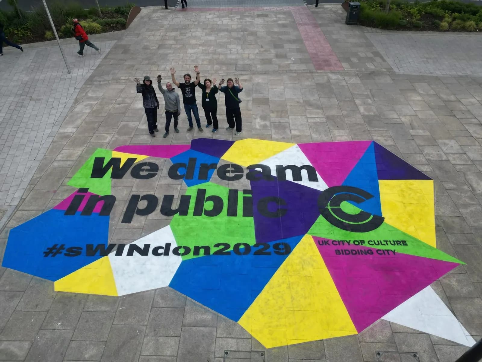 A group of people are waving at the camera from above a colorful ground mural that reads "We dream in public #sWINdon2029" and "UK City of Culture Bidding City".