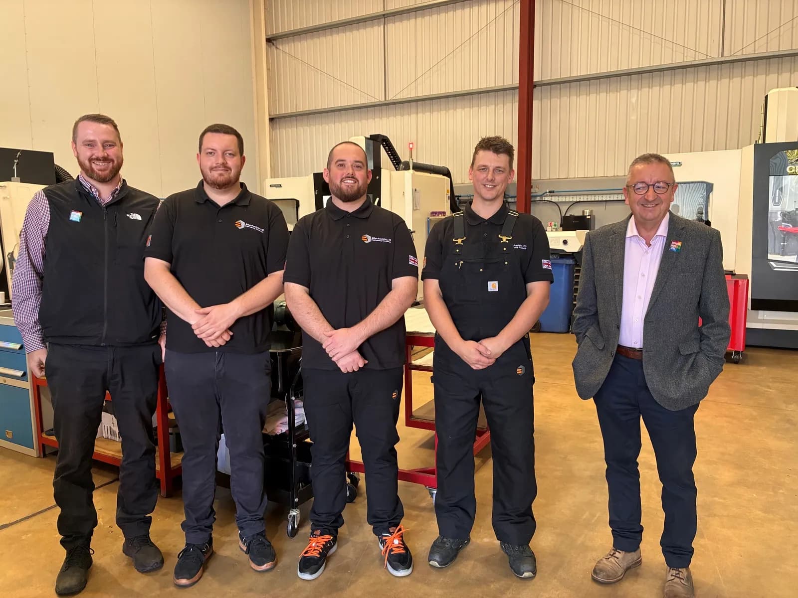 Five men are standing in a row and smiling at the camera inside an industrial building.