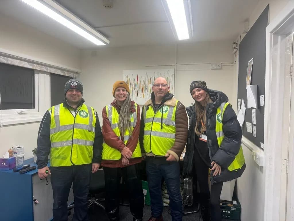 Four individuals wearing high-visibility vests and casual clothing stand smiling in an indoor office setting.