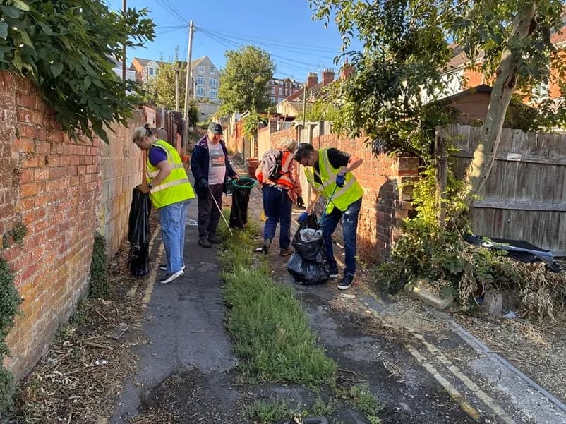 A group of people in high-visibility vests are picking up litter along a narrow, overgrown alleyway between a brick wall and a wooden fence.