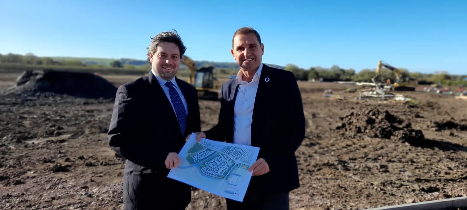 Two men in suits stand smiling in a construction site holding a blueprint, with excavators in the background.