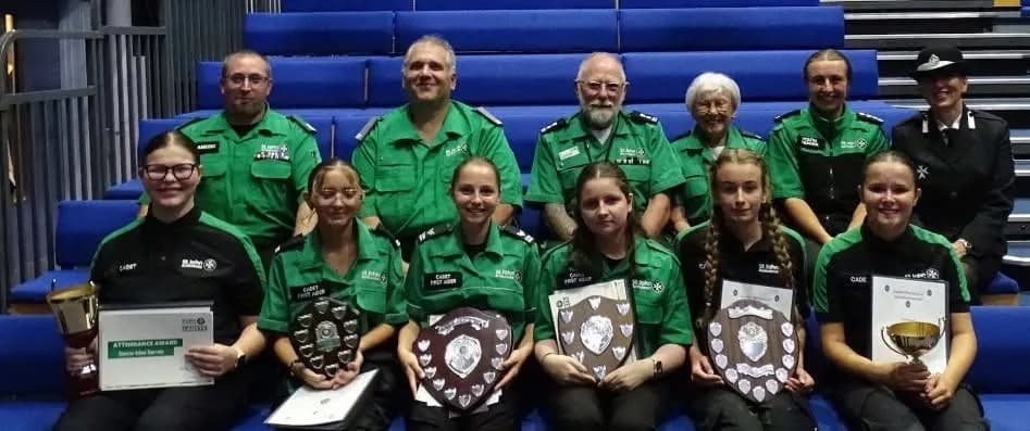 A group of adults and teenagers in green and black uniforms pose together, with several holding trophies and certificates, against a backdrop of blue stadium seating.