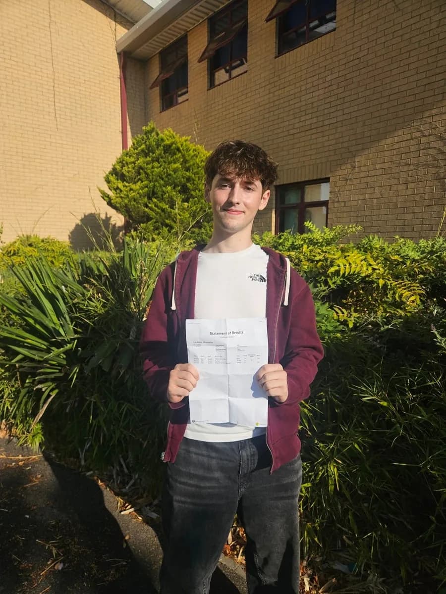 A young man with curly brown hair, wearing a white t-shirt and a maroon zip-up hoodie, smiles while holding up a "Statement of Results" paper with both hands in front of a brick building and lush green bushes.